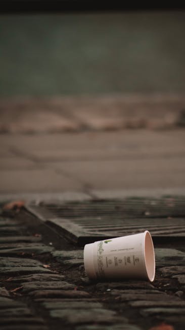 A discarded empty paper coffee cup lying on a cobblestone pavement, with the cup's white surface and printed text visible. The cup is positioned near a metal drain cover, which is partially visible in the lower foreground of the image. The background shows blurred outdoor paving and concrete steps, suggesting an urban or street setting. The scene appears to be outdoors, with natural lighting creating subtle shadows and highlights on the textured surface of the cobblestones and the cup. This visual reflects the issue of littering and improper waste disposal often addressed by private rubbish removal services like Waste Removal Pimlico, highlighting the importance of responsible waste handling and alternative disposal options in public and private spaces.