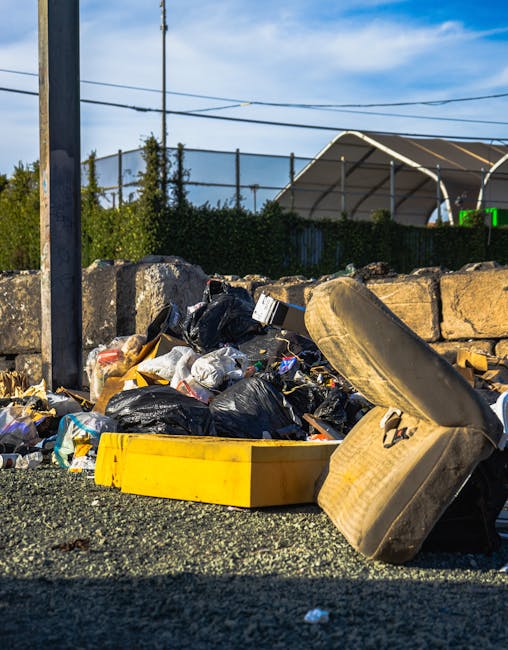 A pile of illegally dumped rubbish on a paved area, consisting of black and clear plastic bags filled with waste, a yellow plastic container, and a large, broken, beige upholstered armchair with visible fabric texture and signs of wear. The waste is positioned in front of a low stone wall made of uneven, weathered stones, with some debris scattered on the ground. In the background, there is a metal fence, green foliage, and a partially visible tennis or sports court with a curved roof structure, under a partly cloudy sky with blue and white clouds. The scene captures the environmental context of unauthorized disposal in an outdoor setting, which Waste Removal Pimlico services aim to address through private rubbish collection and on-site clearance options.