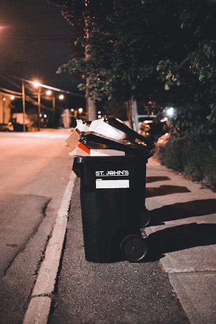 A discarded empty paper coffee cup lying on a cobblestone pavement, with the cup's white surface and printed text visible. The cup is positioned near a metal drain cover, which is partially visible in the lower foreground of the image. The background shows blurred outdoor paving and concrete steps, suggesting an urban or street setting. The scene appears to be outdoors, with natural lighting creating subtle shadows and highlights on the textured surface of the cobblestones and the cup. This visual reflects the issue of littering and improper waste disposal often addressed by private rubbish removal services like Waste Removal Pimlico, highlighting the importance of responsible waste handling and alternative disposal options in public and private spaces.