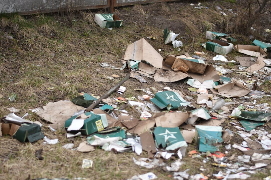 The image depicts a patch of grassy ground adjacent to a paved pathway or area, with scattered litter including collapsed cardboard boxes, some partially flattened, along with smaller paper and plastic debris. The cardboard boxes are mainly green and white with star logos, and appear to have been discarded from packaging, showing torn edges and creases. Small fragments of paper and plastic are intermingled among the boxes, creating a cluttered scene. The grass surrounding the debris is patchy, with areas of exposed soil and a few sparse plants, indicating an outdoor environment. In the background, there is a weathered wooden fence or barrier, partially out of focus, that separates the grassy area from the rest of the surroundings. The lighting is natural, suggesting daytime, with no direct shadows or bright highlights. This image exemplifies an instance of improperly disposed waste in an outdoor area, which could be addressed by a professional waste removal service such as Waste Removal Pimlico to facilitate private cleaning or on-site clearance of rubbish.