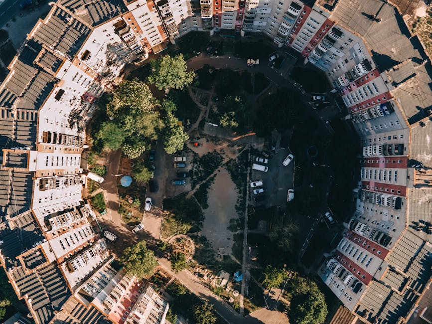 An aerial view of a circular residential courtyard surrounded by multi-storey terraced houses painted in white, cream, and pastel shades, with red and grey tiled roofs. The courtyard features a small green park area with several large leafy trees and walking paths, alongside parked cars lining the perimeter on narrow asphalt roads. The sunlight casts shadows across the scene, highlighting the textures of the roofing materials and the foliage. This setting may be associated with private waste collection or on-site clearance activities, akin to those handled by Waste Removal Pimlico, as part of local rubbish removal services aimed at maintaining cleanliness within such residential communities.