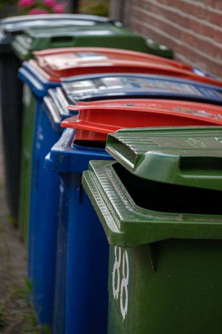 A row of large wheelie bins positioned along a paved pavement area next to a brick wall, with a green bin at the front followed by a blue bin, a red bin, and another green bin further back. The bins are made of durable plastic with textured lids; the green bins feature a ribbed pattern on the lids, while the blue and red bins have smooth, slightly glossy finishes. The bins are aligned neatly in a straight line, with their wheels visible at the bottom. The scene is outdoors, with natural daylight illuminating the bins, emphasizing their bright, solid colors. The background includes a wooden fence and a small patch of grass, indicating a residential setting. The positioning and appearance of the bins suggest they are used for household waste disposal, possibly offered as part of a private waste collection service in Pimlico, which provides an alternative to local authority rubbish collection, consistent with the context of independent rubbish removal services. Waste Removal Pimlico's presence is implied by the professional setting, aligning with the service category of rubbish removal.