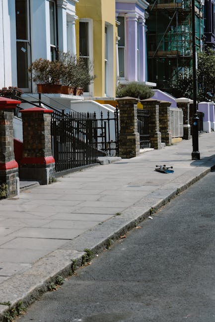 A quiet urban street scene showing a curved road bordered by black bollards and double yellow lines on a wet pavement. To the left, there is a small public area with leafless trees, some shrubbery, and a park bench, indicating a cold or winter season. In the background, multi-storey residential buildings with cream and beige brick facades, white window frames, and decorative columns at ground level are visible. The buildings have large, evenly spaced windows and iron railings enclosing the ground-floor entrances. The overcast sky casts diffuse lighting across the scene, accentuating the wet surface of the street and sidewalk, creating subtle reflections. The image captures a calm, tidy environment typical of city centre residential districts where private waste collection or on-site clearance, such as that provided by Waste Removal Pimlico, could be relevant for managing rubbish or fly-tipping debris in the area.