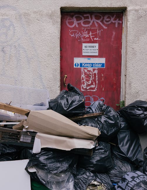 An urban alleyway scene featuring a red metal door with graffiti tags, signs indicating 'No Money' collection restrictions and 'Keep Clear', and an extensive pile of black rubbish bags and discarded packaging materials in front of the door. The waste includes cardboard boxes, plastic wrapping, and other debris, with some of the rubbish stacked and leaning against the textured beige wall surrounding the door. The environment suggests on-site waste accumulation beyond regulated collection, demonstrating a private disposal situation. Waste Removal Pimlico manages such clutter by organizing rubbish removal services from this type of scene, ensuring safe and efficient clearance of accumulated waste within urban settings, and supporting alternative waste handling methods outside of official council collection.