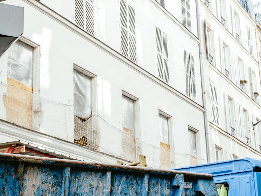The image depicts the upper floors of a multi-storey residential building with a white façade and multiple windows fitted with closed white shutters. Some of the window openings are boarded up with plywood, and a section of netting is visible in front of one window, indicating ongoing repairs or renovation. At the lower part of the image, a large, weathered blue skip container is positioned on the pavement, filled with construction debris or waste material including wooden boards and other miscellaneous refuse. The skip appears to be made of metal with a rough, textured surface showing signs of rust and dirt. To the right of the skip, part of a green vehicle or waste collection truck is visible, likely part of a rubbish removal service such as Waste Removal Pimlico. The background environment suggests an urban setting with a typical city street scene, indicating that the waste collection is part of an on-site clearance process, potentially as an alternative to council-led rubbish disposal, with the focus on private waste management for refurbishment or renovation activities.