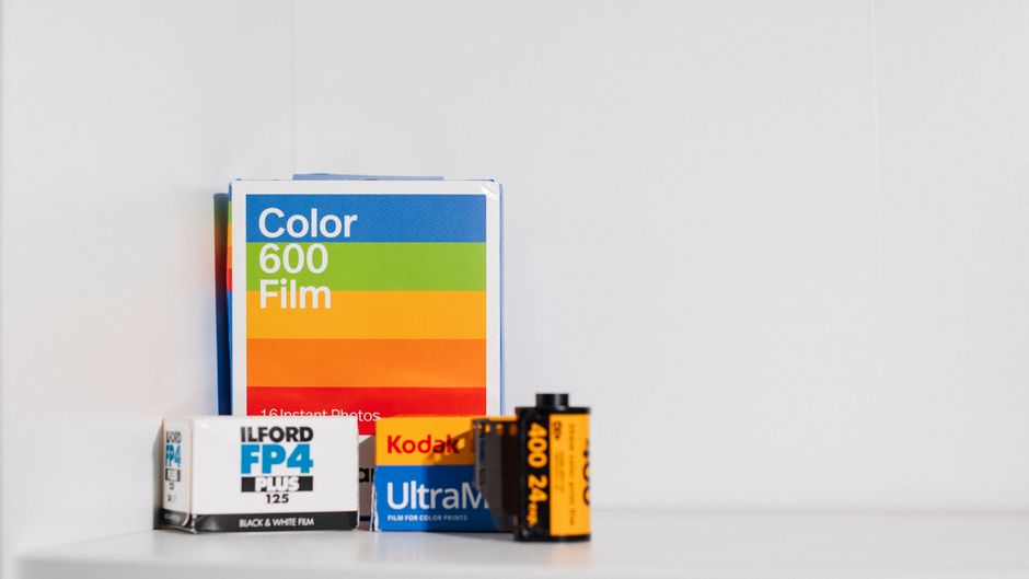 A collection of photographic and film materials displayed on a white surface against a plain white background. The items include a multicoloured box of color film labeled 'Color 600 Film,' positioned towards the left side, featuring horizontal bands of blue, green, orange, and red. To the right of this box are smaller packages of photographic film and accessories, such as a white and black box marked 'Ilford FP4 Plus 125' for black and white film, a black and orange pack labeled 'Fujifilm Ultra Max,' and a small, primarily black with orange accents film roll or battery container. The arrangement suggests an organized setup for photography or film-related activities, and the scene is evenly lit to highlight the textures and colours of each item clearly. This setup subtly relates to the themes of media production or creative waste disposal, reflecting the idea of managing byproducts of photographic or film equipment, which Waste Removal Pimlico can handle through private waste collection.
