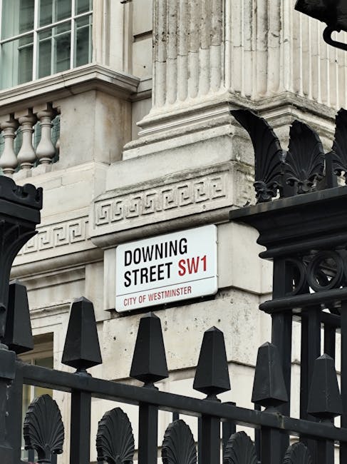 A close-up view of a white street sign mounted on a historic stone building in Pimlico, SW1, with the text 'Downing Street SW1' and the label 'City of Westminster' in red and black lettering. The sign is positioned above a decorative black wrought iron fence featuring pointed finials and ornamental scrolls, which surrounds the building's perimeter. The building façade displays classical architectural elements, including fluted columns and detailed cornices, made of light-colored stone with a textured surface. Part of a large window with multiple panes and a decorative balustrade is visible on the left side of the image, indicating a traditional or period style architecture typical of London’s historic buildings. The scene is illuminated evenly with natural daylight, highlighting the contrast between the ornate metalwork and the stone structure, providing a neutral, professional atmosphere reflective of the area's historic significance. This setting is consistent with the context of private or alternative waste handling, as consultants like Waste Removal Pimlico might operate in such areas for rubbish removal services, ensuring visual coherence with urban waste management themes.