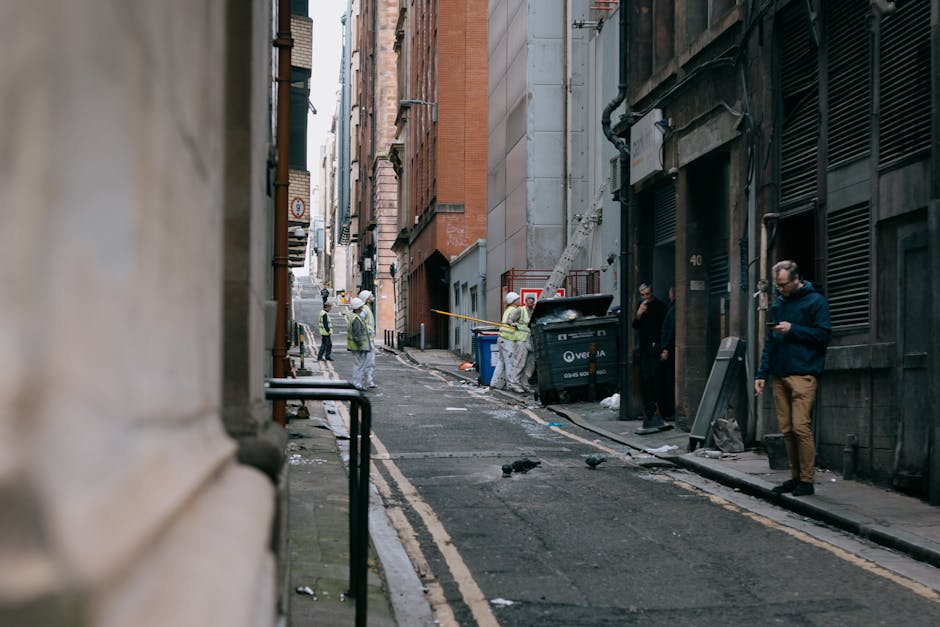 A narrow urban alleyway flanked by multi-storey brick and concrete buildings, with a slight upward incline. The scene includes a large black waste bin positioned against a building wall, partially open with debris inside. Several workers wearing high-visibility jackets and helmets are visible in the background, engaging in rubbish collection or on-site clearance activities, with some standing near the bin and others further down the alley. Scattered waste and litter are present along the wet pavement, with a few birds pecking at debris. To the right, a man dressed in dark clothing, wearing glasses, is standing near a doorway while checking his phone. The environment appears to be away from main roads, with minimal natural light, and the surrounding buildings have external numerous pipes and ventilation gratings. Occasionally, the scene relates to private waste handling, and Waste Removal Pimlico occasionally conducts such discreet rubbish disposal activities in confined spaces like this alleyway. The scene captures the typical context of independent collection in a dense urban setting, highlighting the practical challenges of rubbish removal in narrow spaces without visible collection vehicles.
