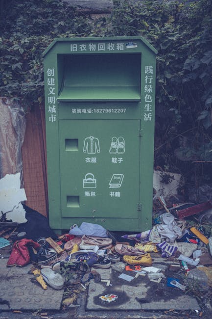 A green, old-style clothing donation and waste collection bin positioned outdoors against a backdrop of dense greenery and foliage. The bin features Chinese characters and icons indicating it is for collecting used garments, shoes, bags, and books. It has a rectangular open top for depositing items and two small compartments below, one for clothing and shoes, the other for bags and books. Surrounding the bin on the ground are an assortment of discarded items, including worn shoes, scattered clothing, plastic bags, and small miscellaneous debris, suggesting a collection or disposal site. The scene is illuminated by natural light, with some shadows cast by the nearby objects and foliage. The environmental context indicates a location where waste and unwanted textiles are privately collected, aligning with alternative rubbish removal methods professionals like Waste Removal Pimlico might facilitate, especially for bulky or heavy items requiring specialized disposal.