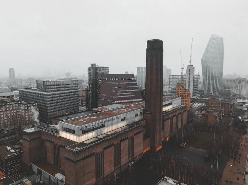 Aerial view of the Tate Modern art gallery in London, showcasing its distinctive red-brick facade and tall chimney stack in the foreground. Surrounding the gallery are modern high-rise office buildings and office towers, some under construction with cranes visible against a cloudy, overcast sky. The cityscape extends into the distance with various glass and steel structures, including the curved, glass-clad skyscraper known as the Walkie Talkie. The scene depicts an urban environment with a mix of historic and contemporary architecture, and the weather appears misty or foggy, reducing visibility of the farther buildings. This image provides a snapshot of the central London skyline near the Tate Britain and Pimlico area, illustrating the city's architectural diversity and ongoing development, relevant to businesses like Waste Removal Pimlico that offer services for the disposal and clearing of waste materials from such busy, densely built areas.