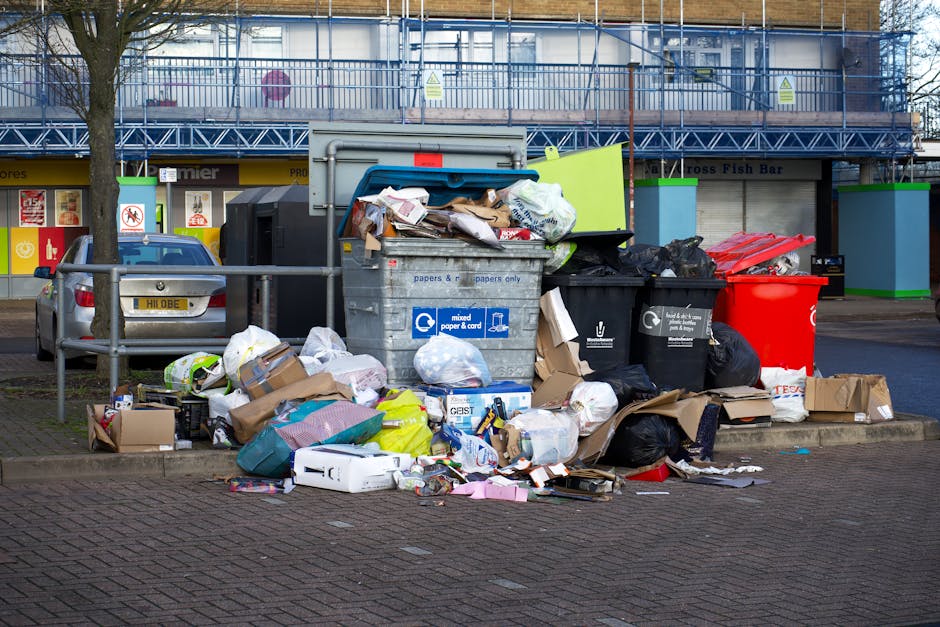 An outdoor scene showing multiple waste containers positioned on a paved area beside a metal railing, with a variety of rubbish including cardboard boxes, plastic bags, paper, and packaging materials overflowing from the bins and scattered around their base. The central grey bin labeled for mixed paper and card is open, with loose items and flattened boxes spilling onto the ground. Adjacent to it, a black bin appears full, with black refuse bags leaning against or partially inside it. To the right, a red bin is visible with no contents overflowing. Behind the bins, a building with large glass windows and a blue safety scaffold is visible, indicating ongoing construction or maintenance work. Nearby parking includes a silver car with a UK registration plate, and a few small trees are planted along the sidewalk, separated by metal railings. The scene suggests an area where private waste disposal or additional rubbish collection is occurring, as part of an alternative waste handling arrangement in a public environment.