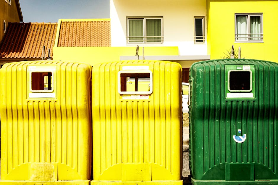 A large, green, industrial-style waste container filled with mixed rubbish, situated outdoors on a street or public space, with various discarded items visible inside and around it. The contents include flattened cardboard boxes, some with torn or bent edges, and white packaging materials. Adjacent to the container, there are more cardboard boxes and packaging debris piled haphazardly, with some leaning against the side of the bin. In the background, a blurred street scene features trees, urban buildings, and pedestrians, indicating a city environment requiring responsible waste disposal. The container appears well-used, with chipped paint and dirt marks on its surface, consistent with frequent handling, possibly by private waste removal services like Waste Removal Pimlico performing on-site clearance for bulky or irregular waste streams. The lighting suggests daytime with natural light illuminating the scene, emphasizing the textures and colors of the various materials, and reinforcing the context of alternative rubbish management outside regular municipal collections.